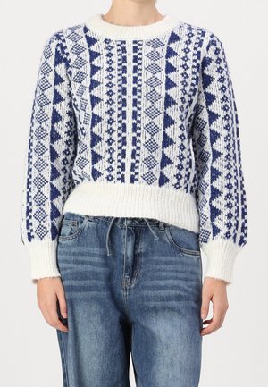 Pull blanc en maille avec des motifs géométriques bleus et des manches bouffantes, porté avec un jean en denim bleu.