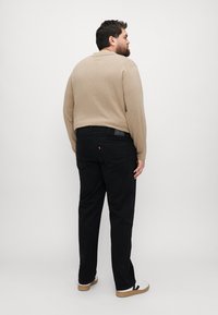 Beige langermet polo-skjorte og svarte jeans med avslappet passform. Mørke bukser har en baklomme med etikett. Nøytral bakgrunn.