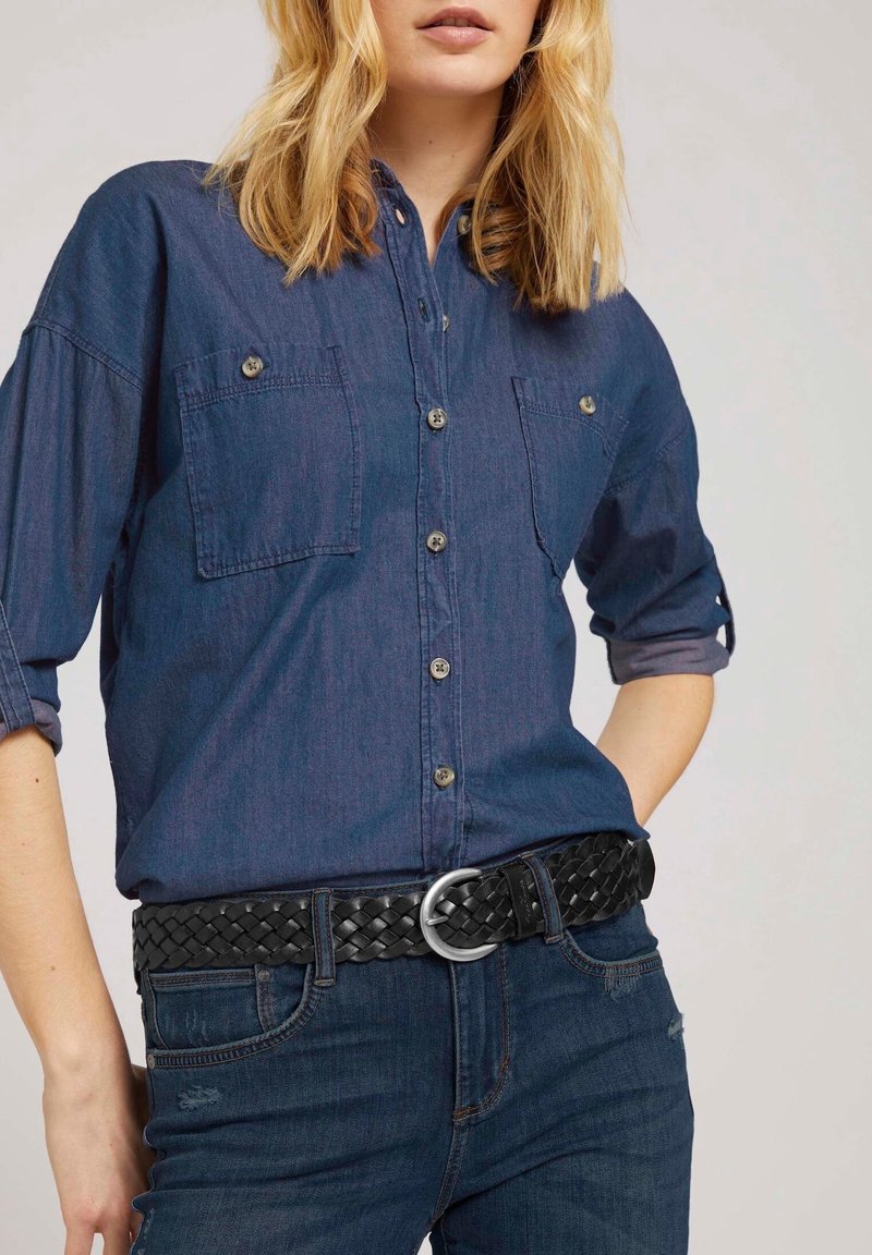 Femme portant une chemise en denim foncé à manches longues avec deux poches poitrine et une ceinture tissée noire par-dessus un jean foncé.