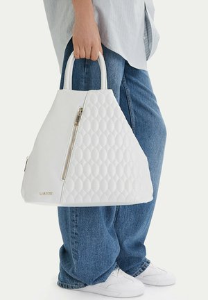 Persona sosteniendo un gran bolso acolchado blanco con cremallera dorada, vistiendo jeans azules holgados, zapatillas blancas y una camisa ligera a rayas.