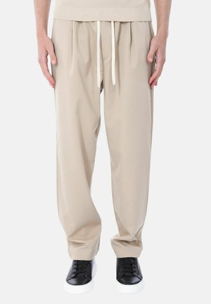 Pantaloni beige dal taglio comodo con pieghe frontali e coulisse bianca, abbinati a sneakers nere con suole bianche.