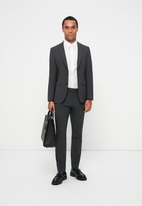 Completo grigio su misura con revers a punta, camicia bianca, scarpe nere e una borsa in pelle nera. Tessuto liscio e design aderente.