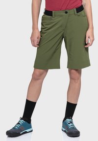 Grüne Shorts mit geradem Schnitt, versehen mit einem schwarzen Taillenbund und einer vorderen Reißverschlusstasche. Kombiniert mit blauen und schwarzen Wanderschuhen sowie schwarzen Socken.