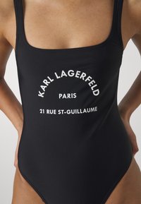 Črni enodelni kopalke s kvadratnim izrezom. Ima bele napise: "KARL LAGERFELD PARIS 21 RUE ST-GUILLAUME." Gladka tekstura in prilegajoča oblika.
