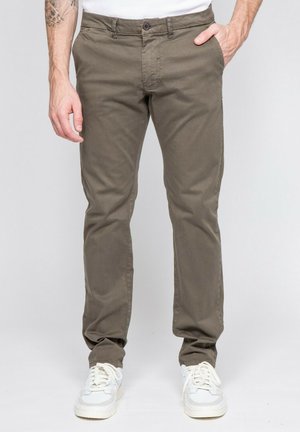 Homme portant un pantalon slim kaki et des baskets blanches, debout avec une main dans la poche sur un fond uni.