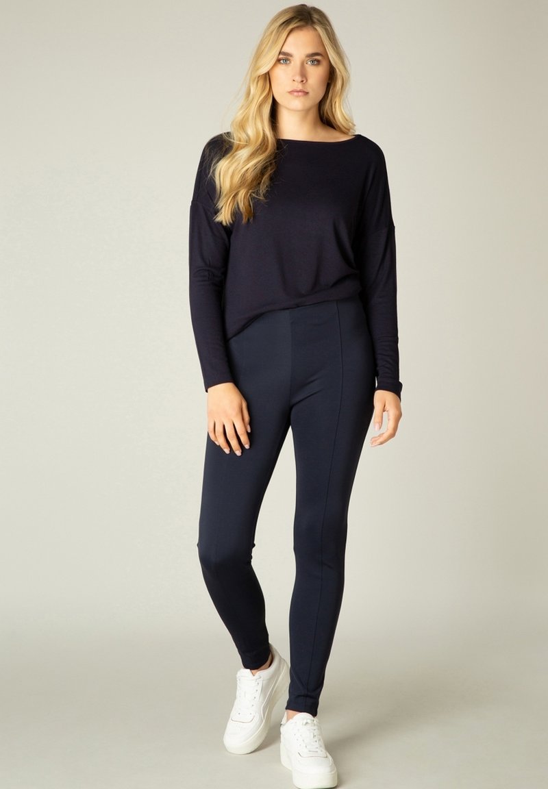 Marineblauwe longsleeve top gecombineerd met marineblauwe nauwsluitende legging. Gladde stof zonder zichtbare patronen. Model draagt witte sneakers.
