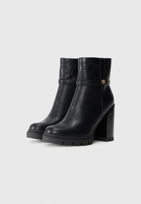 Botas de tobillo de cuero negro con un upper texturizado que presenta un patrón de logotipo grabado, tacón alto y robusto, y suela gruesa.