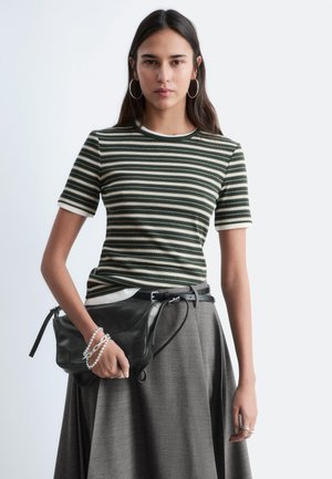 Ung kvinde iført en grøn- og hvidstribet kortærmet skjorte, grå nederdel, øreringe i ringform, som holder en sort lædertaske med perlearmbånd.