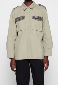 Khaki utility jacket with chest pockets featuring patterned fabric, subtle shoulder accents, long sleeves, and a button-down front.