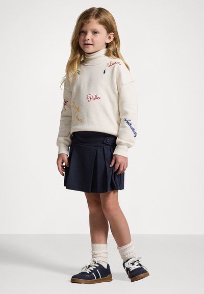 Sweater branco de mangas compridas com texto bordado multicolorido, combinado com uma saia plissada azul marinho. Sapatos escuros e meias claras completam o look.