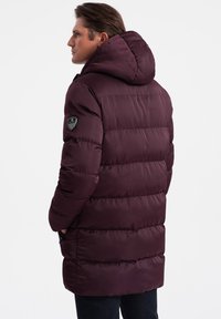 Giacca a vento bordeaux con cappuccio, design trapuntato e logo sulla manica sinistra. Presenta una texture liscia e lucida e una lunghezza fino al ginocchio.