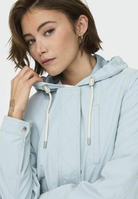 Jeune femme aux cheveux courts et bruns portant une veste à capuche bleu clair, touchant le cordon de la capuche près du visage, avec des boucles d'oreilles et un tatouage sur la main.