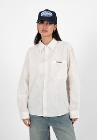 Femme portant une casquette de baseball bleu marine avec un logo blanc, une chemise boutonnée blanche à fines rayures avec poche et logo, et un jean bleu clair sur fond blanc.