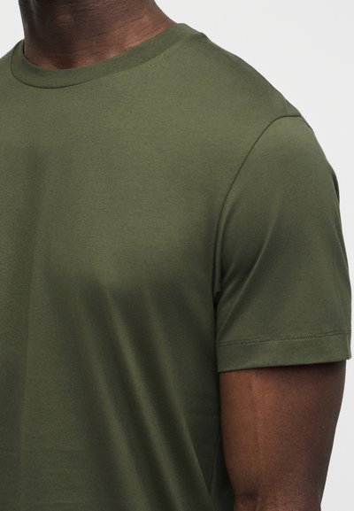 Εγγύς πλάνο ενός ατόμου που φοράει ένα απλό πράσινο ελιάς κοντομάνικο μπλουζάκι με στρογγυλή λαιμόκοψη, μπροστά σε ουδέτερο φόντο.