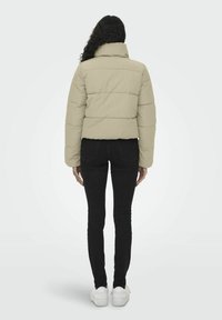 Kort, brun pufferjakke med høy krage og horisontale sømmer, båret over svarte bukser og hvite joggesko; sett bakfra.