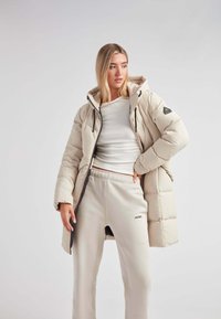 Abrigo acolchado en beige claro, con capucha, cierre frontal y detalles en negro. Combinado con pantalones de chándal beige claro que muestran un logo.