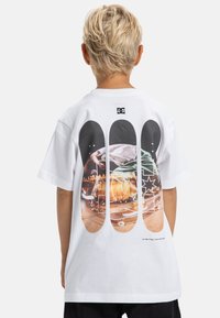 Camiseta de algodón blanca con un diseño gráfico que presenta imágenes de skate y logotipos en la parte posterior. Manga corta, cuello redondo, ajuste casual.