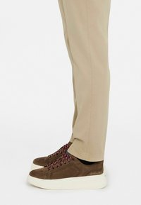 Zapatillas de suede marrón con cordones burdeos y suelas blancas texturizadas, combinadas con pantalones beige ajustados en una postura de pie.