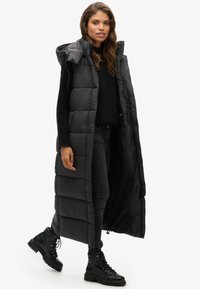 Schwarze, gepolsterte lange Weste mit Kapuze, mit gestepptem Muster und vertikalen Nähten. Kombiniert mit einem schwarzen Pullover und enganliegenden Jeans.