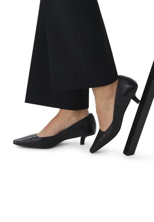 Escarpins en cuir noir avec un bout pointu et un petit talon. Design élégant avec une texture lisse. Portés avec un pantalon noir large à côté d'un pied de chaise.