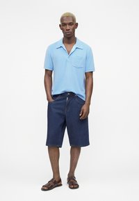 Homme aux cheveux blonds courts portant un polo bleu clair à manches courtes, un short en jean foncé longueur genou, et des sandales marron ouvertes aux orteils, debout.