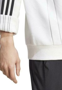 Sweatshirt de sport blanc présentant des détails à trois bandes noires sur les manches, avec des poignets et un ourlet côtelés, associé à un pantalon noir. Texture de tissu lisse.