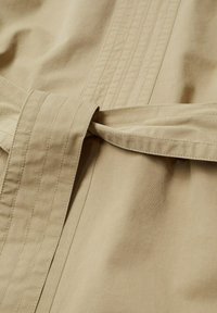 Gros plan sur une ceinture en tissu beige nouée par un simple nœud avec des coutures verticales visibles sur un matériau en coton lisse.