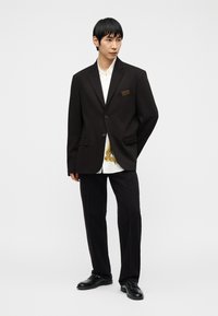 Νεαρός άνδρας που φοράει μαύρο μεγάλο σακάκι, λευκό πουκάμισο με χρυσή εκτύπωση, μαύρο παντελόνι και μαύρα παπούτσια, στέκεται μπροστά σε απλό φόντο.