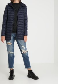 Tmavě modrá prošívaná bunda se zapínáním na zip, prošívaným designem a kapucí, v kombinaci s potrhanými modrými d�žínami a černými šněrovacími botami.