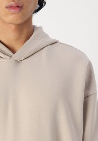 Béžová mikina s jemnou strukturou, s tkaničkou u krku a uvolněným střihem. Obsahuje minimální švy a žádné viditelné vzory nebo kovové prvky.