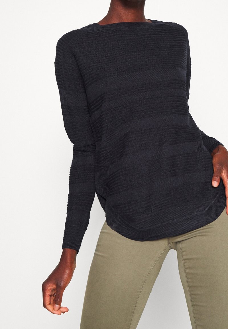 Pull en maille bleu marine foncé avec un motif de rayures horizontales texturées, coupe décontractée, col rond large et manches longues. Associé à un pantalon vert olive.