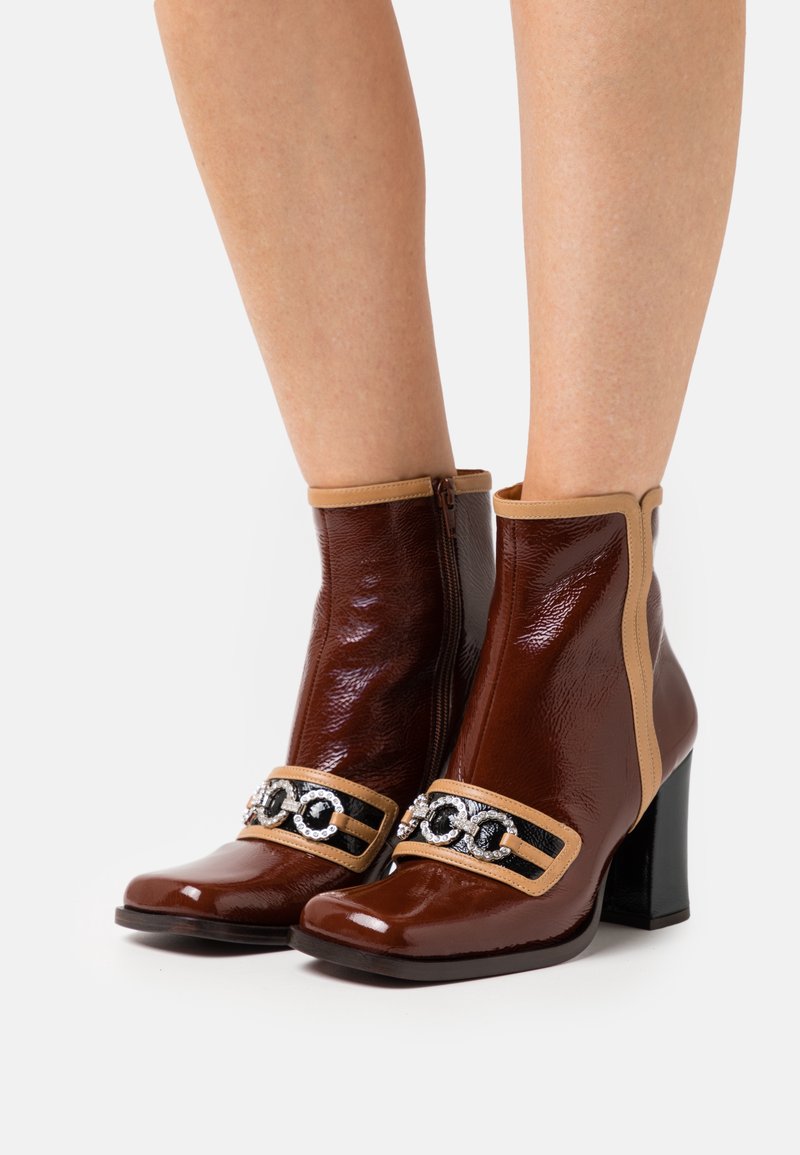 Bottes cheville en cuir verni marron avec un accent beige, fermeture éclair et détail décoratif de chaîne traversant le devant ; design à talon bloc.