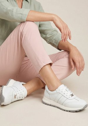 Person iført lyserøde bukser og lysegrøn skjorte sidder med benene i kors og viser hvide sneakers med grå såler på en beige baggrund.