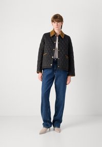 Jachetă neagră matlasată cu guler din suede maro, accesorii aurii și cusături diagonale. Asortată cu un top de culoare deschisă și pantaloni din denim albastri.