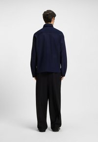Navy shirt with a structured collar and long sleeves, viewed from the back. Paired with loose black trousers, showcasing a relaxed fit.