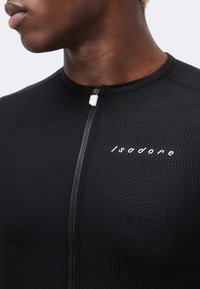 Κοντινό πλάνο ενός ατόμου που φοράει μαύρο υφασμάτινο τοπ με φερμουάρ και λευκό λογότυπο "Isadore" στο στήθος.