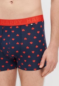 Tmavomodré boxerky s červeným vzorom srdiečok a kontrastným červeným elastickým pásom s vyrazeným slovom "HAPPY". Hladká textúra a priliehavý strih.