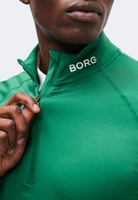 Το χέρι ενός άντρα τραβάει το φερμουάρ ενός πράσινου μπουφάν με την επιγραφή «BORG» στον γιακά, δείχνοντας μέρος του προσώπου και του λαιμού του.