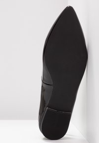 Chaussure en cuir verni noir à bout pointu vue par la semelle, taille 37, appuyée contre un coin blanc.