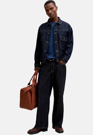 Mann trägt dunkle Jeansjacke und Jeans, blaues Hemd, hält mit einer Hand eine große braune Ledertasche, steht vor weißem Hintergrund.