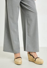 Pantalones de pierna ancha de rayas grises combinados con sandalias de cuña marrón que cuentan con una correa negra acentuada con herrajes dorados y un detalle de yute trenzado.