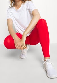 Biała koszulka z subtelnym logo w połączeniu z obcisłymi, czerwonymi legginsami. Model ma na sobie białe buty sportowe. Neutralne tło.