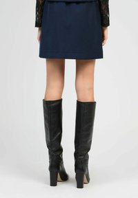 Femme portant des bottes montantes en cuir noir à talons et une jupe bleu marine, vue de dos sur un fond uni.