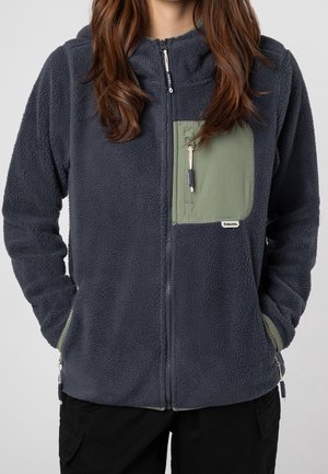 Femme portant une veste en polaire gris foncé avec une poche poitrine zippée verte et des poches latérales bordées de vert, mains dans les poches latérales.