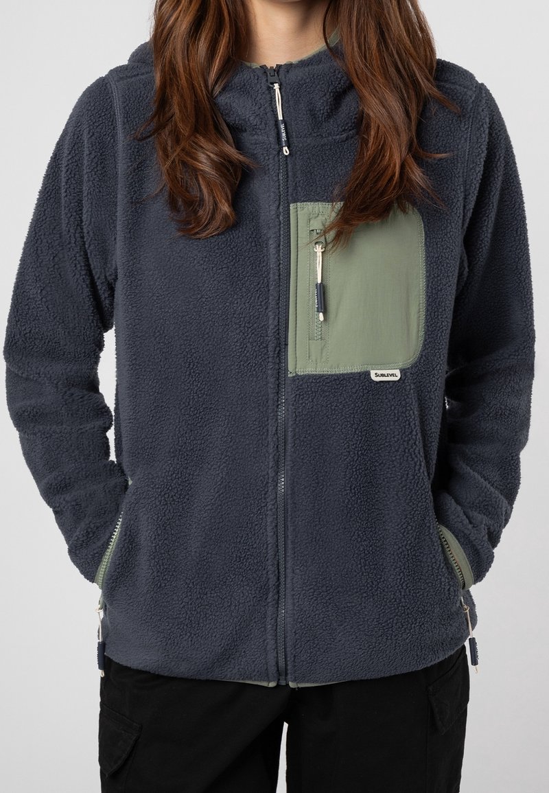 Femme portant une veste en polaire gris foncé avec une poche poitrine zippée verte et des poches latérales bordées de vert, mains dans les poches latérales.
