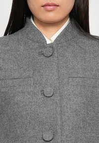 Közelkép egy személyről, aki magas galléros szürke gyapjúkabátot visel, textillel borított gombokkal egy fehér ing felett.