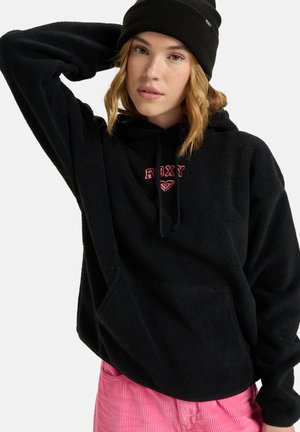 Haut en molleton noir avec poche kangourou, arborant le logo "ROXY" rose. Porté avec un pantalon en velours côtelé rose et un bonnet noir.