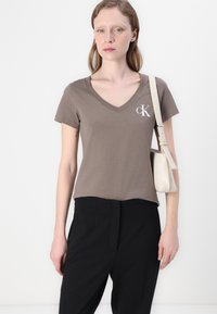 Ruda, prigludusi V formos marškinėliai iš minkštos medvilnės, su balta Calvin Klein logo kairėje krūtinės pusėje. Suderinti su juodomis kelnėmis ir šviesia rankine.