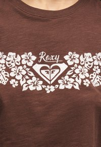 Καφέ ύφασμα με λευκά λουλουδάτα σχέδια, λογότυπο σε σχήμα καρδιάς και τη λέξη «Roxy» τοποθετημένη στο κέντρο πάνω από το λογότυπο.