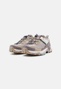 Ζευγάρι πεζοπορικών παπουτσιών σε μπεζ και μοβ με παχιά, στιβαρή σόλα, ελαστικά κορδόνια και λευκό ρυθμιζόμενο ιμάντα πάνω σε λευκό φόντο.